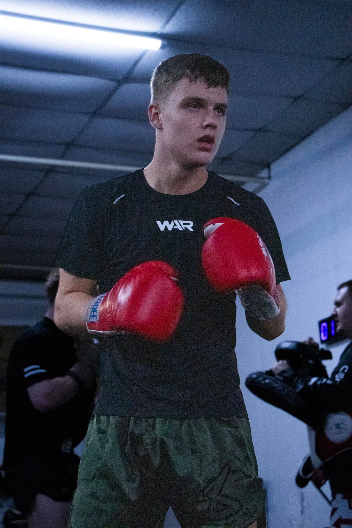 A young man in a boxing stance wearing a black athletic t-shirt with 'WAR' printed on the chest, red boxing gloves, and green camouflage boxing shorts inside a gym.