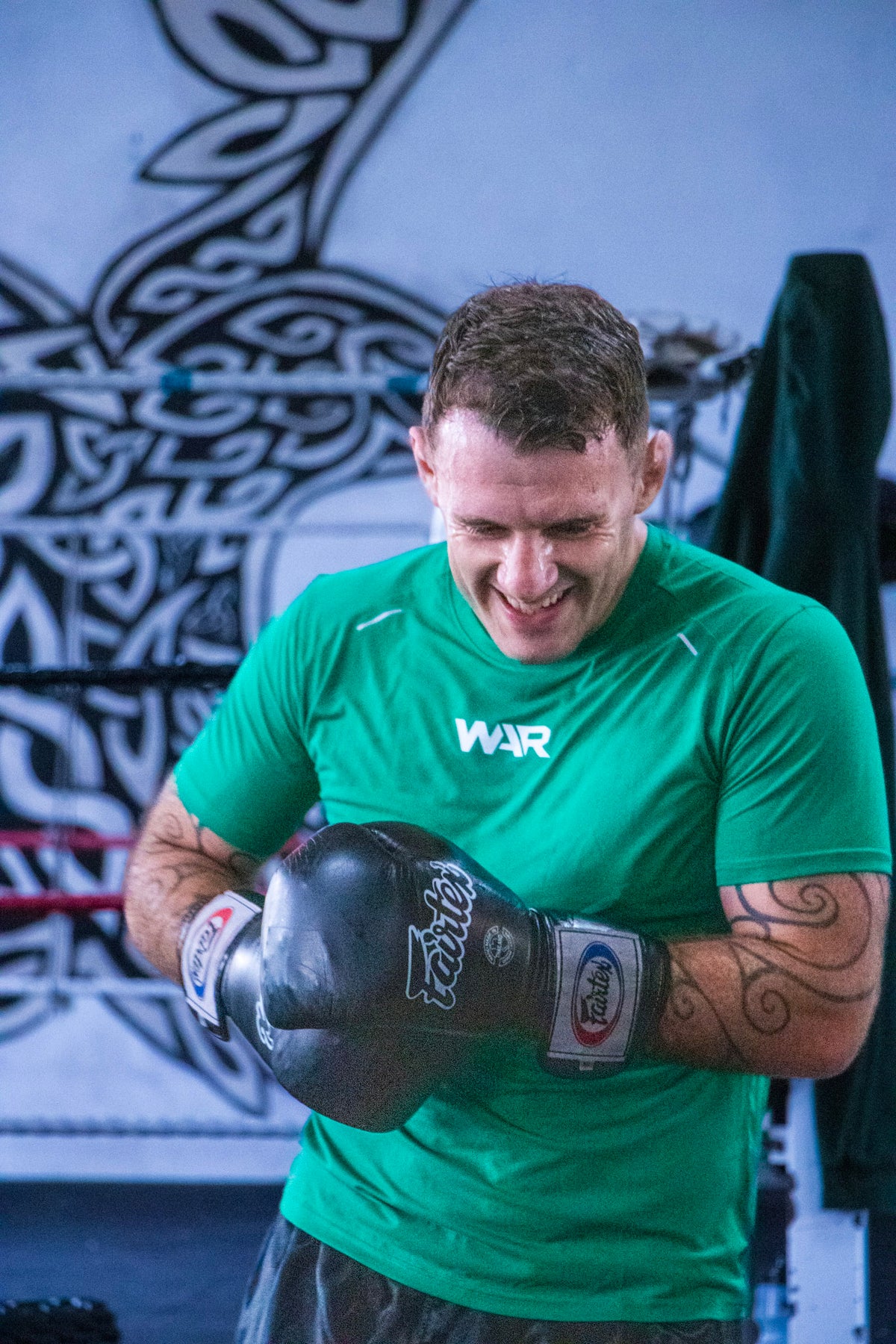 A man in a green athletic t-shirt with a white 'WAR' logo and black boxing gloves, smiling while looking down in a gym setting with a Celtic-style wall mural.