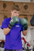 A young man in a boxing stance wearing green boxing gloves and a purple athletic t-shirt with 'WAR' printed on the chest, set against a gym background with stacked cardboard boxes.