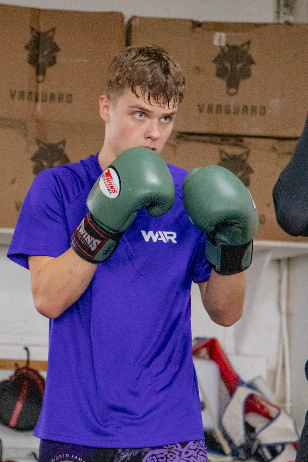 A young man in a boxing stance wearing green boxing gloves and a purple athletic t-shirt with 'WAR' printed on the chest, set against a gym background with stacked cardboard boxes.